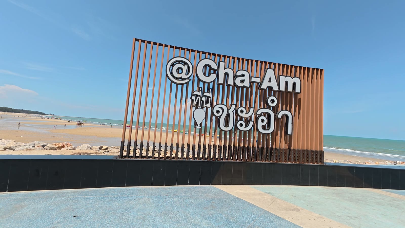 Das Ortsschild @Cha-Am aus Holzlamellen an der Strandpromenade, dahinter der Sandstrand und der Golf von Thailand
