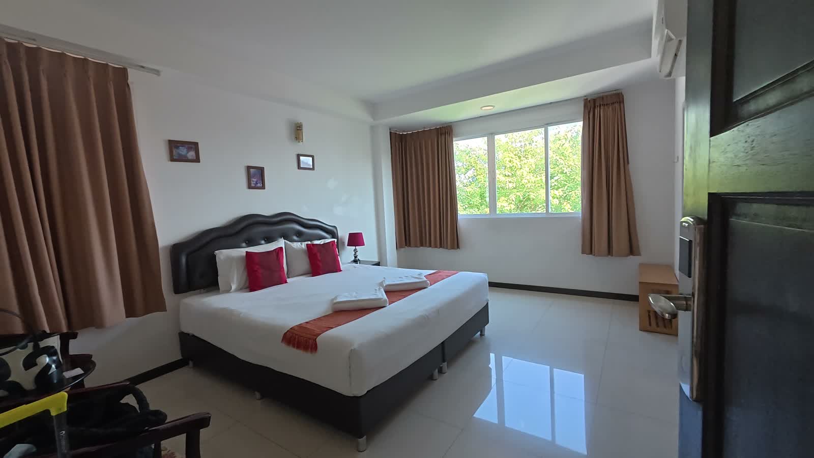 Hotelzimmer im TH Beach Hotel in Hua Hin: Doppelbett mit roten Kissen, helle Vorhaenge, Fliesenboden und Fenster mit Blick ins Gruene