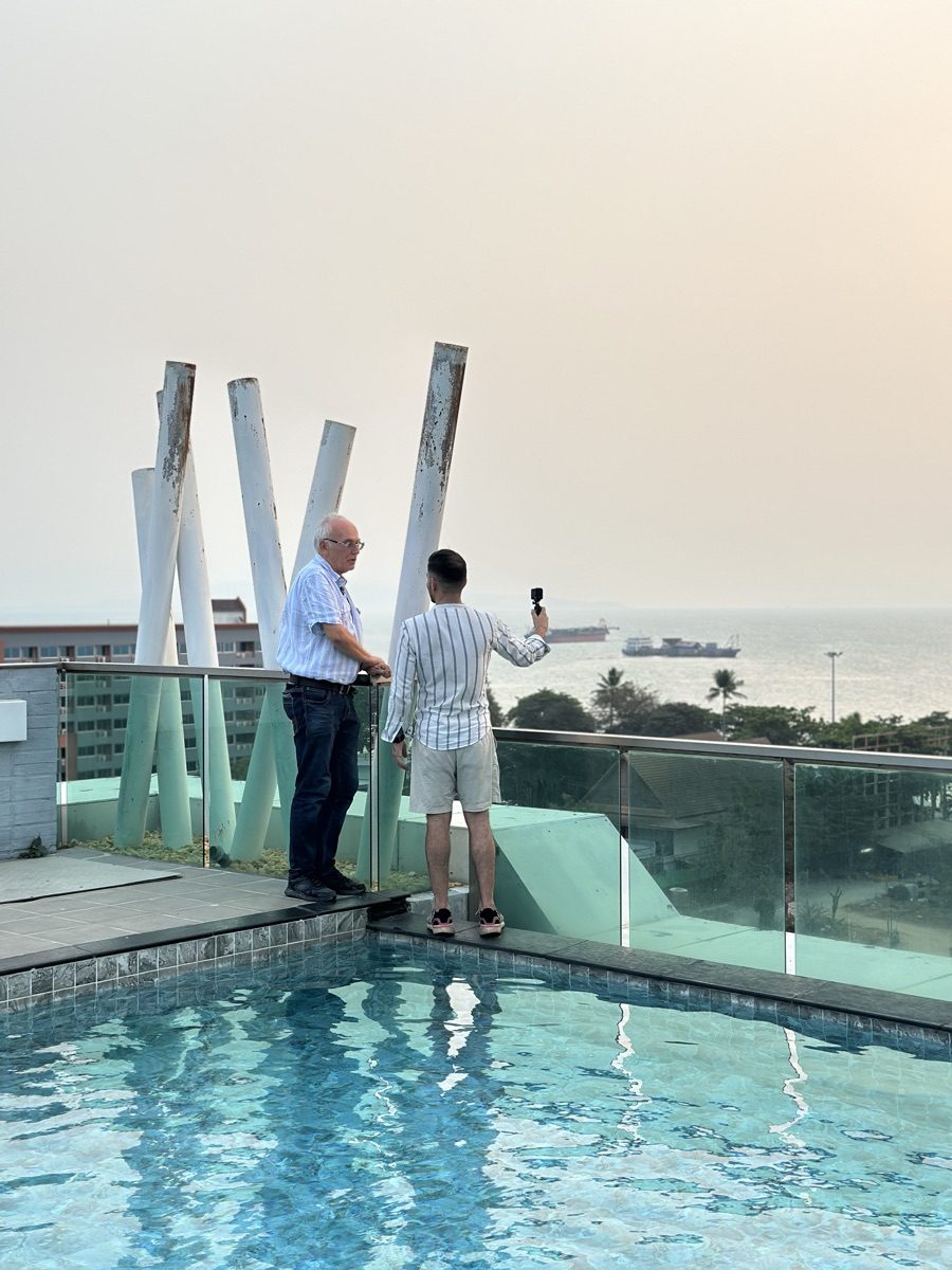 Nihat filmt mit einer kleinen Kamera einen älteren Auswanderer auf der Dachterrasse einer Wohnanlage, dahinter der Pool und die Bucht von Pattaya.